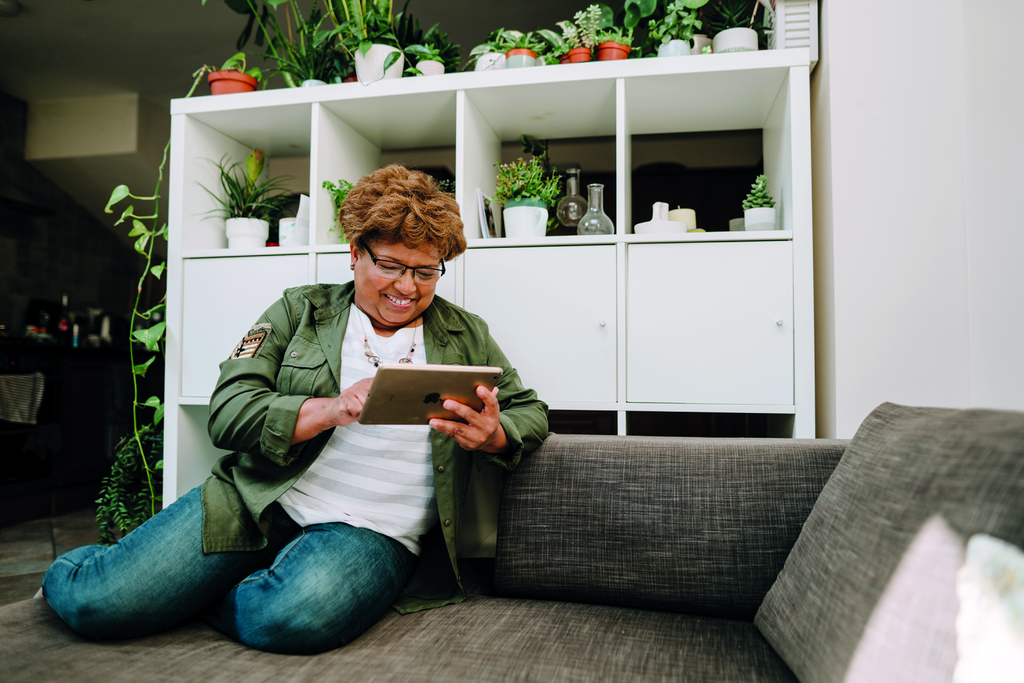 ältere Frau sitzt am Sofa und macht etwas an ihrem Tablet